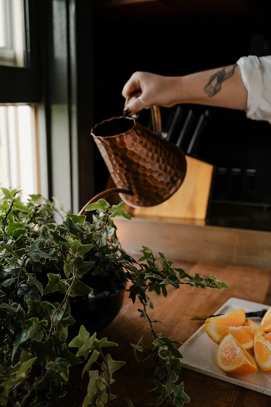 Person pouring water from a hammered copper pitcher onto a potted ivy plant on a wooden table, with a plate of sliced oranges nearby.
