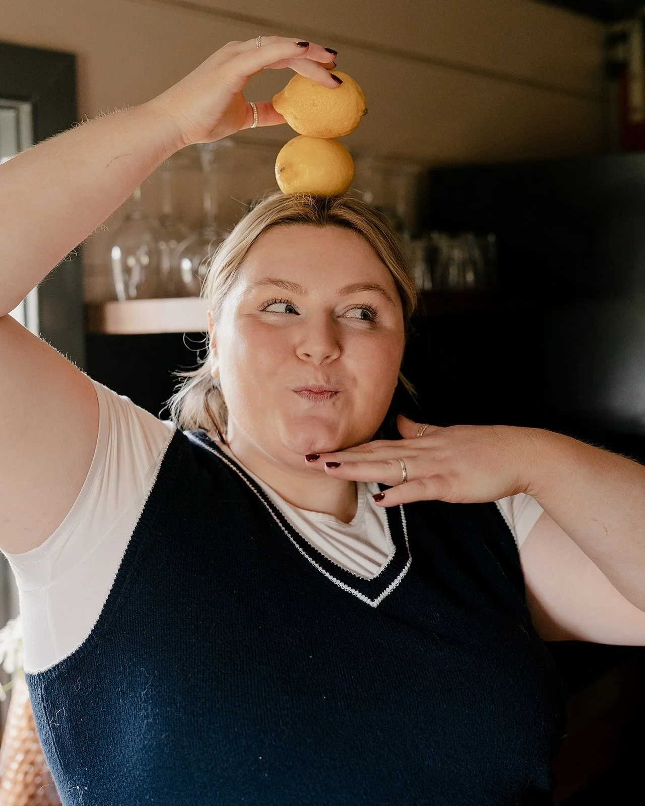 An ADHD dietitian making a playful pose with her hand under her chin and a raised arm holding two lemons balanced on her head in a cozy indoor setting.