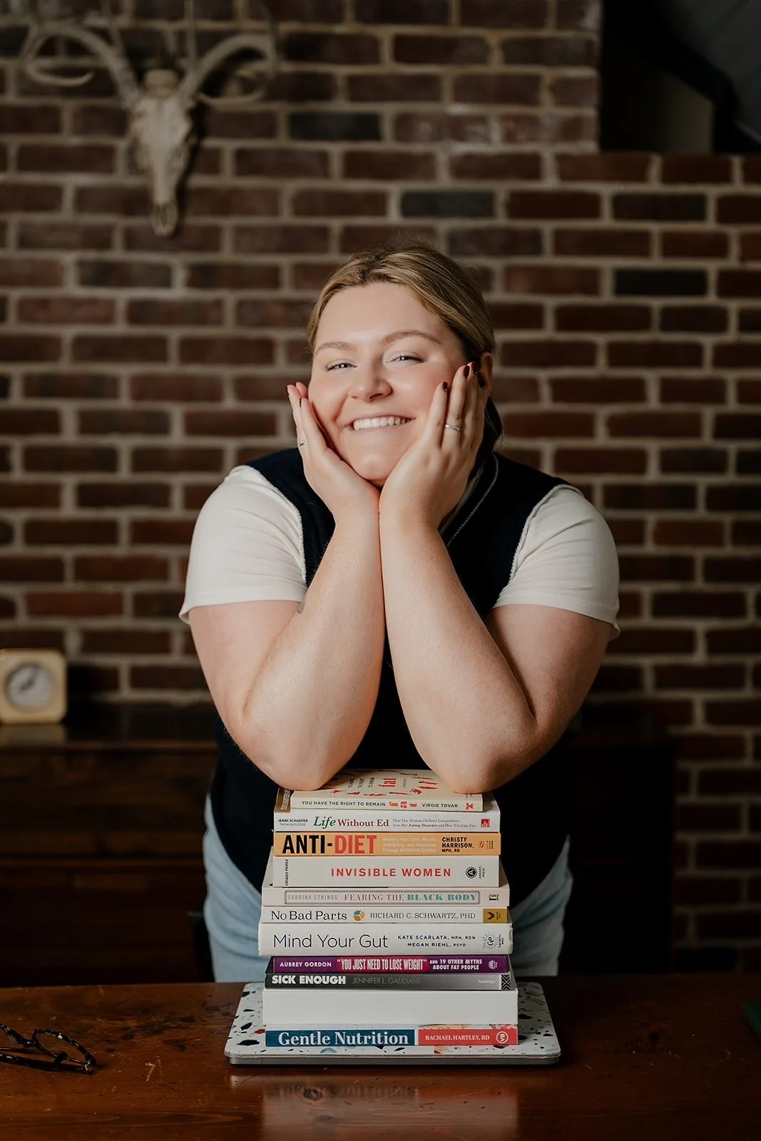 Woman dietitian nutritionist with blonde hair smiling and leaning on her hands, resting on a stack of books on a wooden table, against a brick wall with a deer skull mounted above.