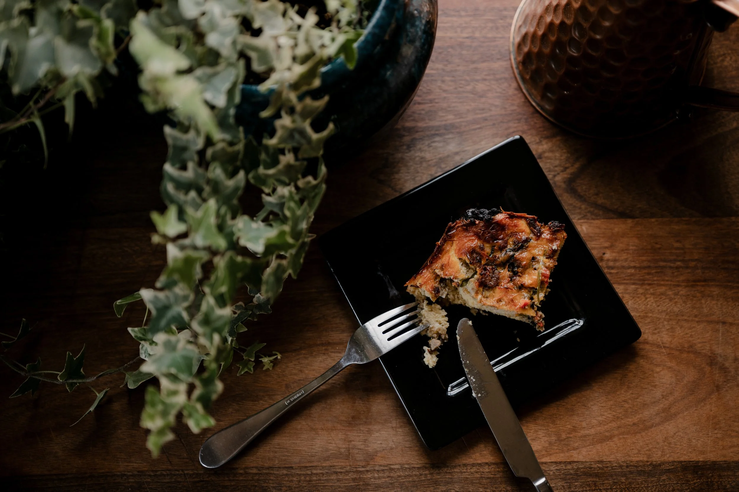 A partially eaten slice of vegetable quiche on a black square plate, with a fork resting on the plate, placed on a wooden table. A potted plant with green leaves is partially visible along the left side of the image. Additionally, there appears to be a container of pepper and a container of dried herbs on the right side of the image.