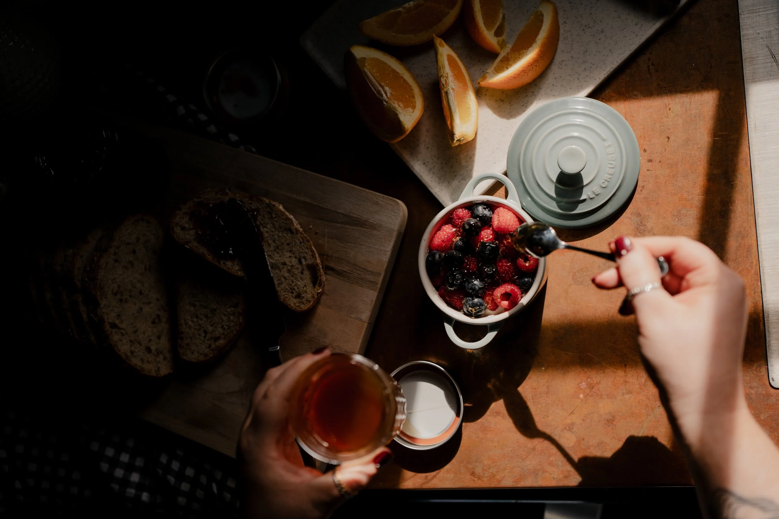 A kitchen countertop with sliced oranges on a cutting board, a cup with mixed berries, a hand holding a spoon, a glass of beverage, slices of bread, and an orange and a wooden surface on the right side.