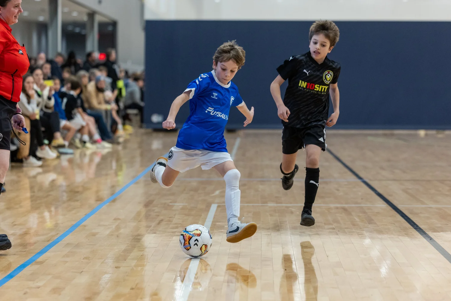 Two young boys playing indoor futsal, chasing a ball on a wooden court, with a crowd of spectators sitting on the side.