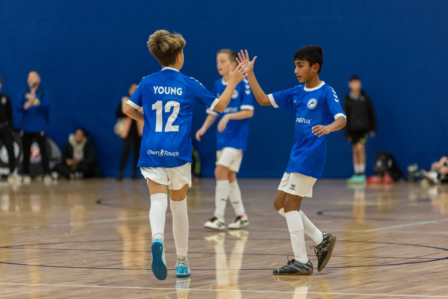 Two young boys in blue soccer uniforms high-five on an indoor court, with other players and spectators blurred in the background.