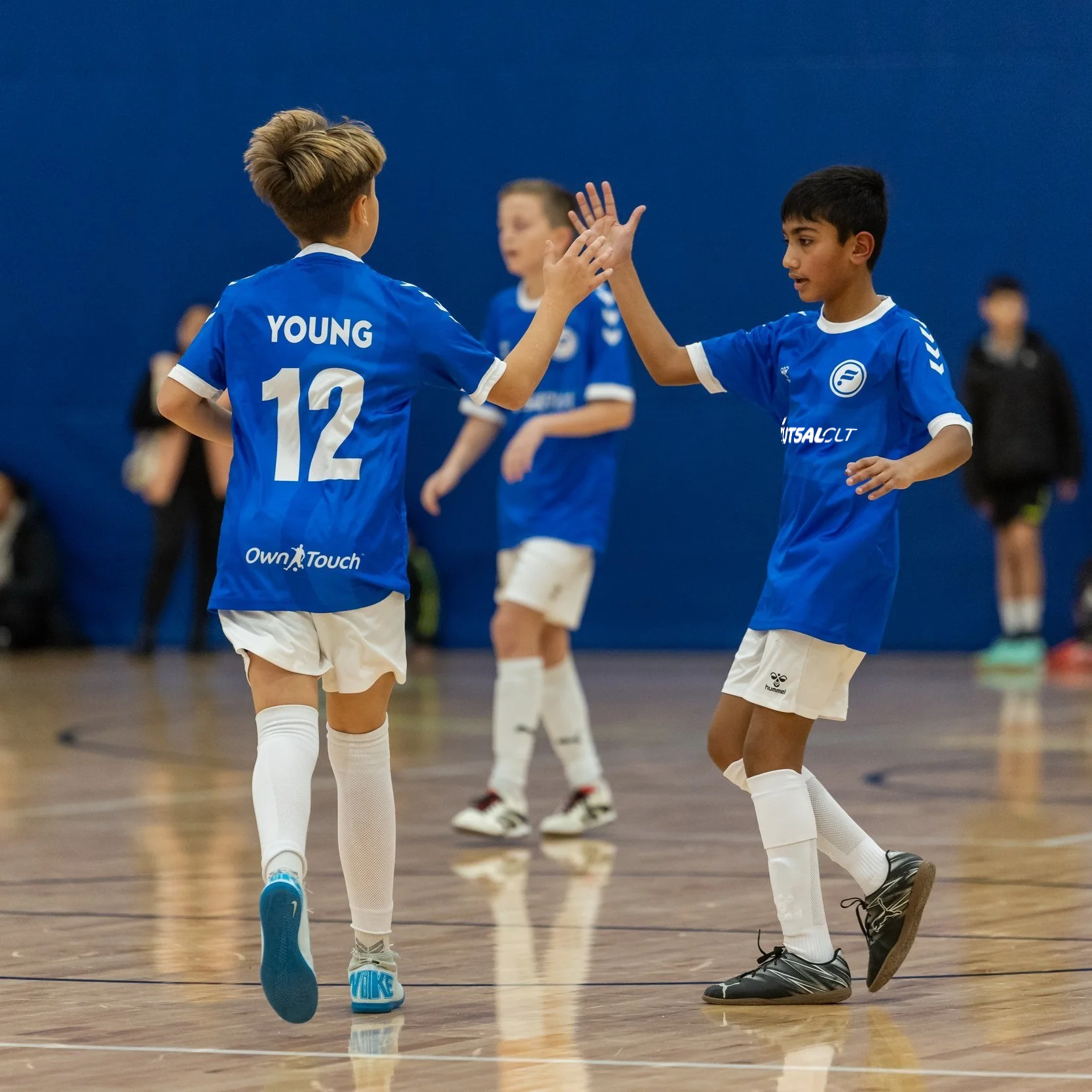 Two young boys in blue charlotte futsal uniforms high-fiving each other in an indoor futsal hall with other children in the background.