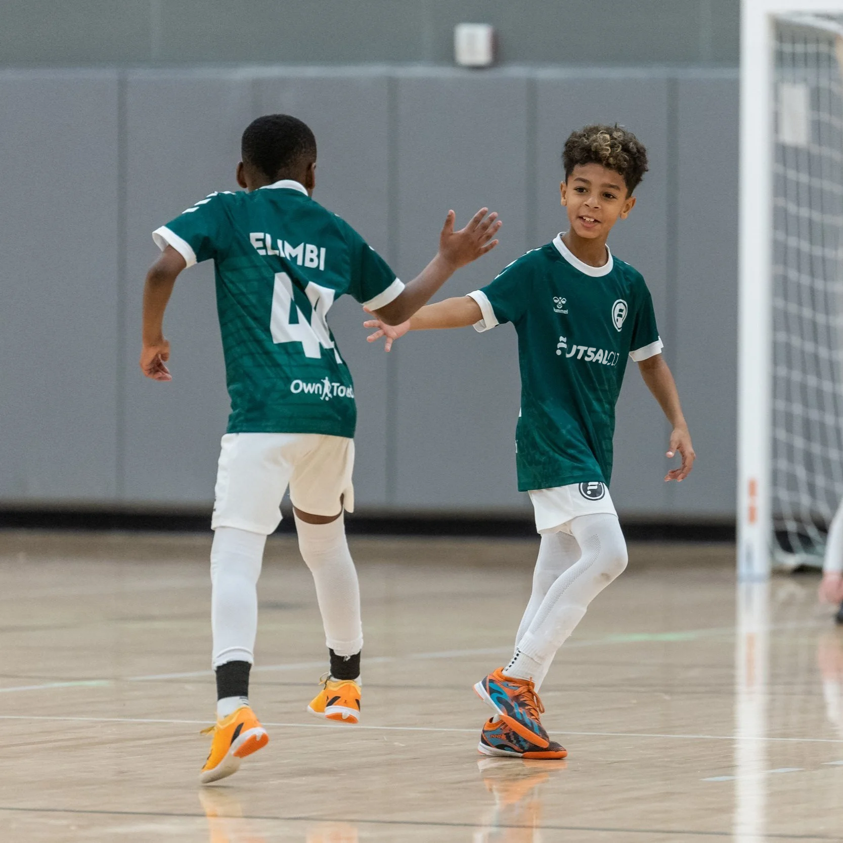 Two young boys wearing green charlotte futsal uniforms celebrating during an indoor futsal game, giving high fives, with a goalpost visible in the background.