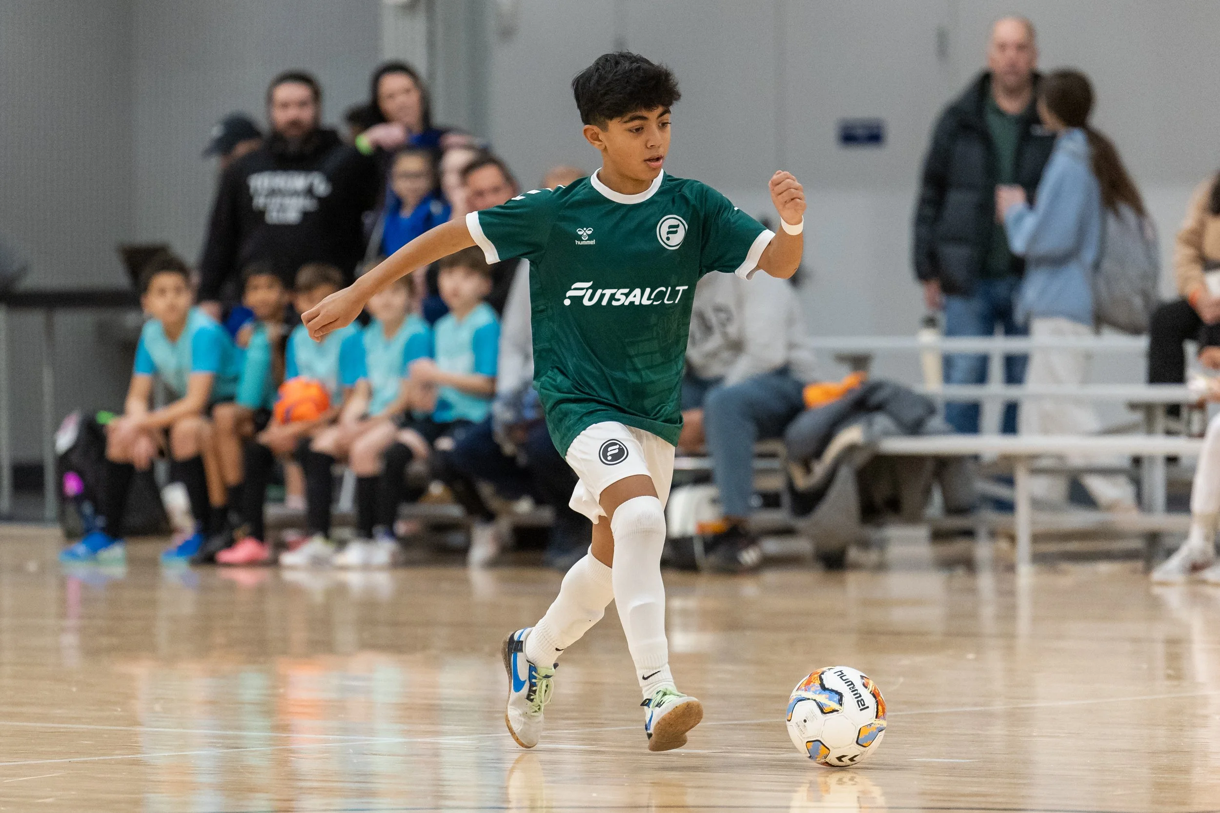 A young boy in a green futsal uniform dribbling a futsal ball during indoor game, with spectators and teammates sitting on benches in the background.