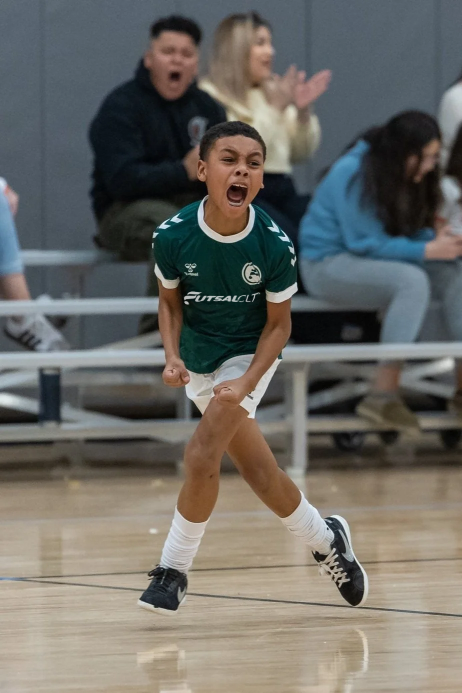 Young boy in green soccer jersey celebrating on indoor court, with spectators in background clapping and cheering.
