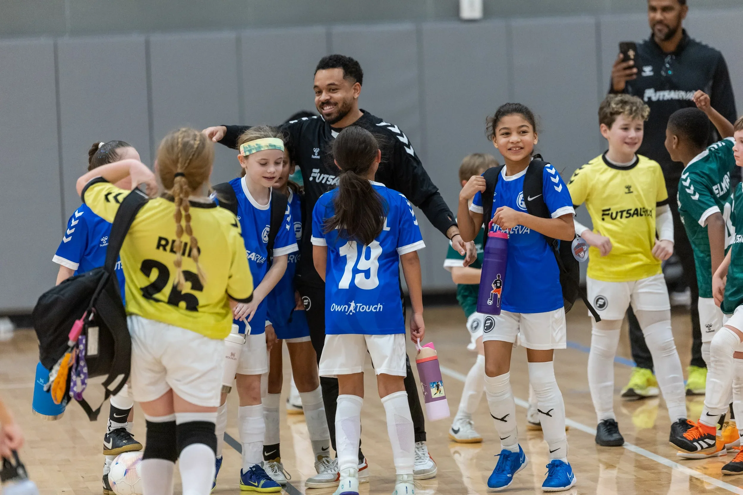 Kids in sports jerseys and shorts gathered in a gym with a coach. The kids are smiling and interacting with each other, holding water bottles and wearing backpacks.