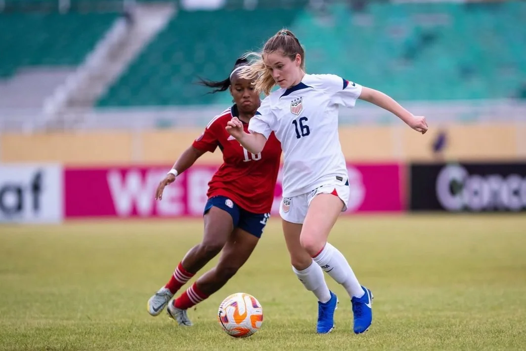 Two female soccer players competing for ball on field, one in white USA jersey and one in red and blue jersey, in stadium setting.