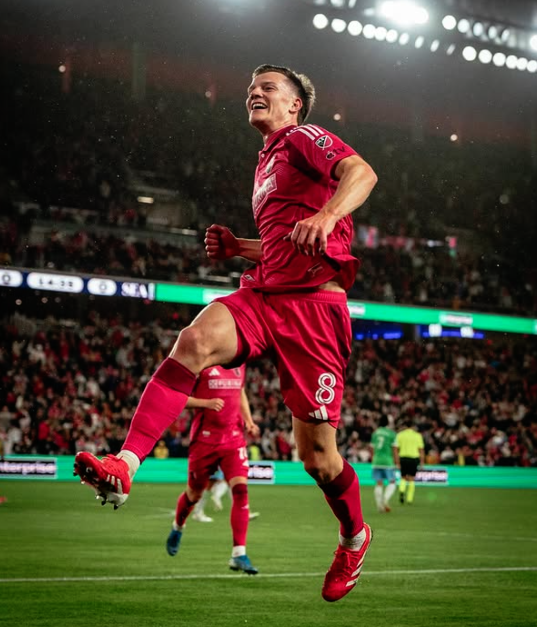A soccer player wearing a red uniform with the number 8 on shorts, celebrating by jumping on the field during a match in a large stadium filled with spectators, under bright stadium lights.