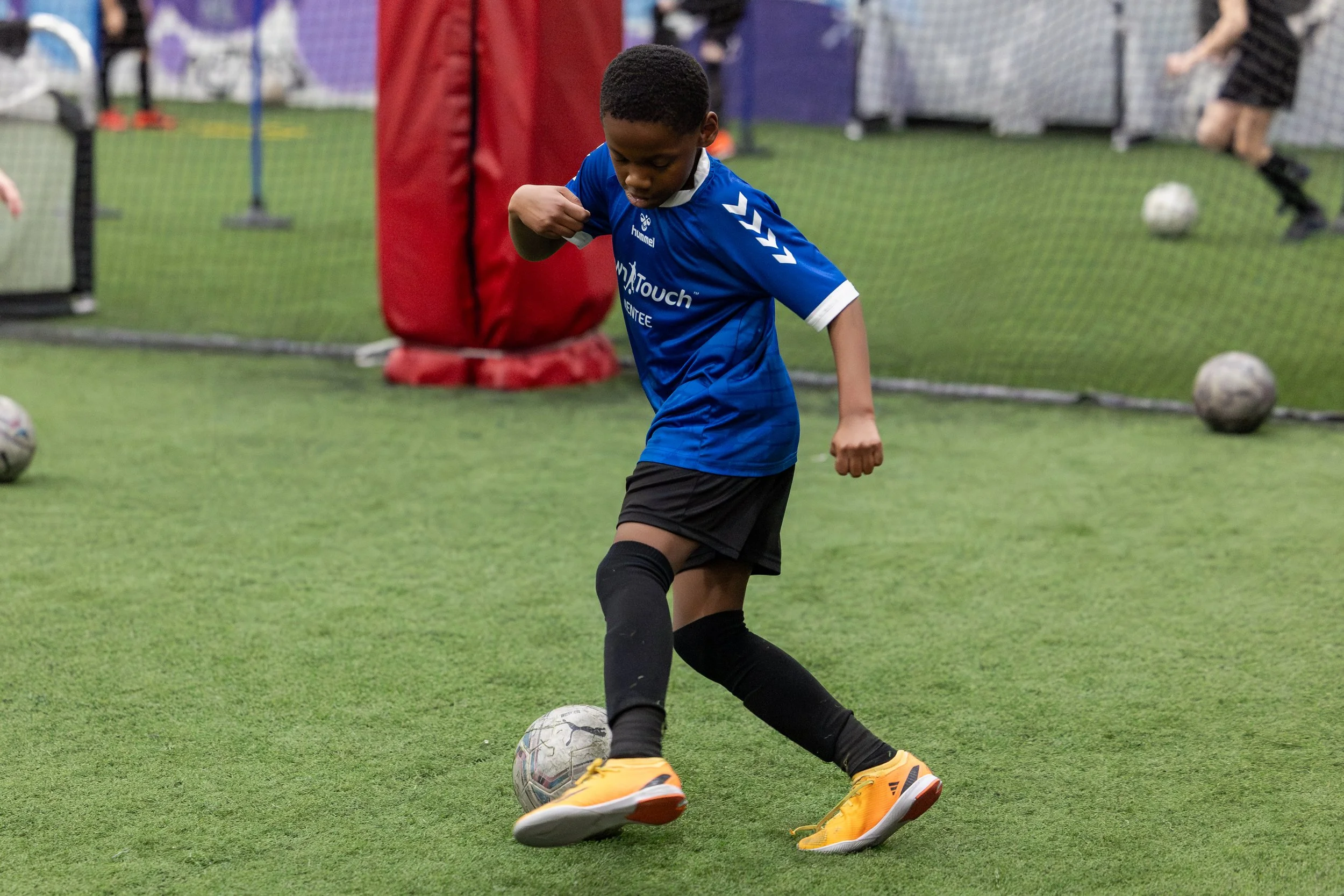 A young boy in a blue sports jersey and black shorts playing soccer indoors. He is kicking a soccer ball with orange and yellow cleats. There are multiple soccer balls on the artificial turf and a netted wall in the background.