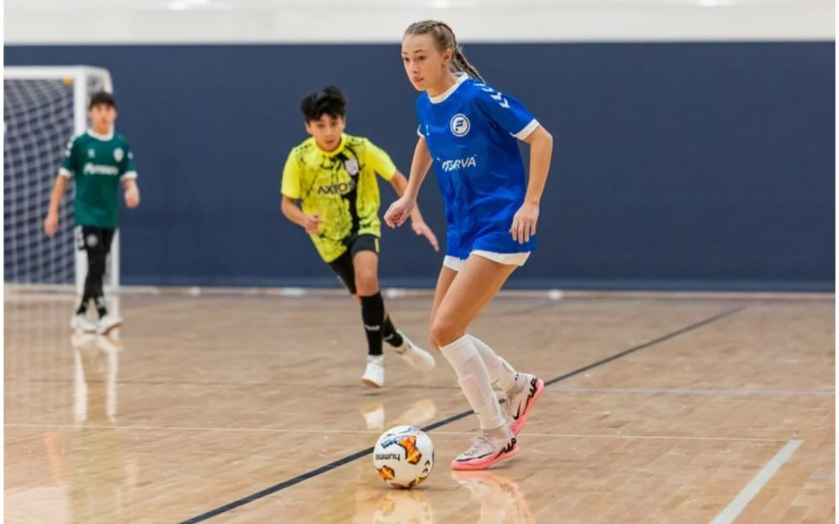 A girl in a blue soccer uniform dribbling a futsal ball on an indoor court, with two boys in the background chasing her.