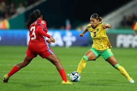Two female soccer players, one in a red uniform and the other in a yellow and green uniform, compete for the ball on a soccer field.