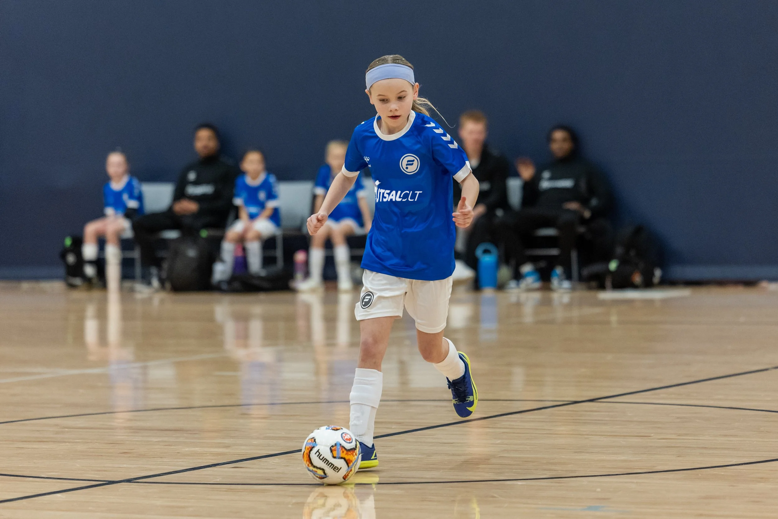 Young girl in blue sports jersey and white shorts playing indoor soccer, dribbling a soccer ball on a wooden court, with teammates and coaches sitting on benches in the background.
