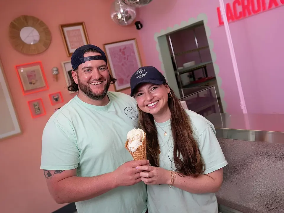 A man and woman smiling inside an ice cream shop, holding a cone with two scoops of ice cream.