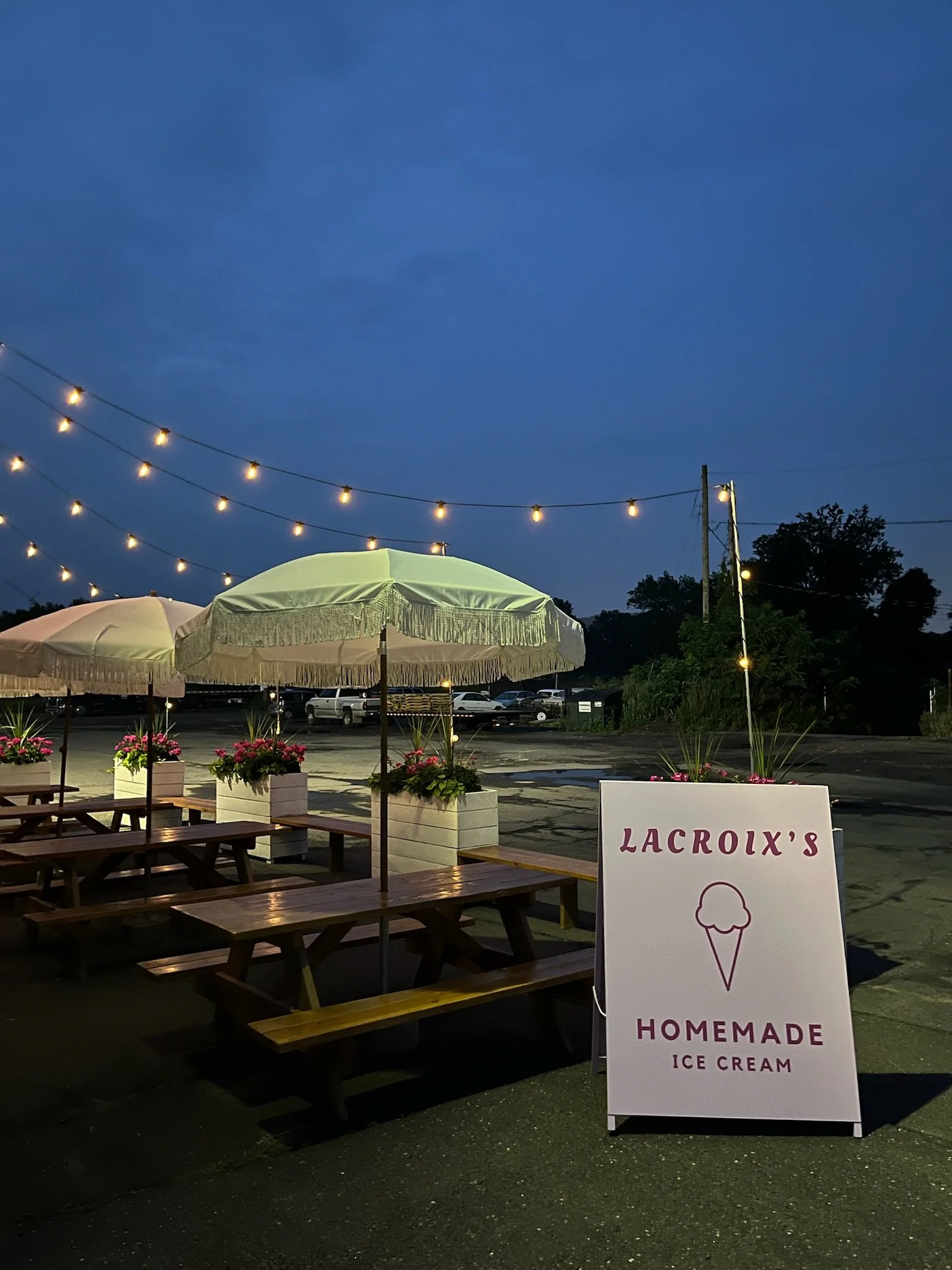 Outdoor evening scene of a homemade ice cream stand with a sign that reads 'Lacroix's Homemade Ice Cream', surrounded by picnic tables and potted flowers, illuminated by string lights against a darkening sky.