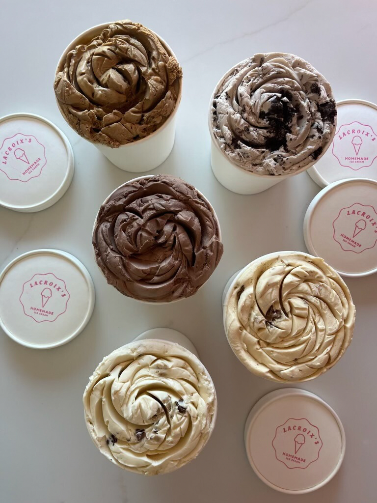 Five cups of assorted ice cream flavors with sprinkles and swirls, along with four empty lids labeled 'Lacroix’s Homemade Ice Cream' on a white surface.