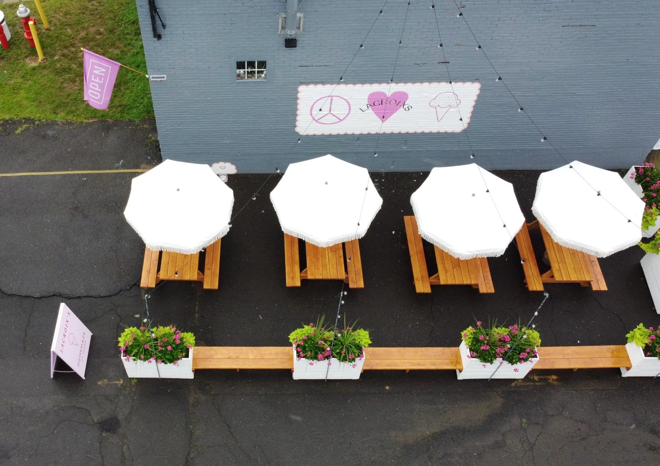 An aerial view of an outdoor seating area with four white umbrellas over wooden tables on a black pavement, decorated with pink and purple flowers in white planters, adjacent to a blue building with a sign reading 'L'ACROPOIS' and a pink 'OPEN' flag.
