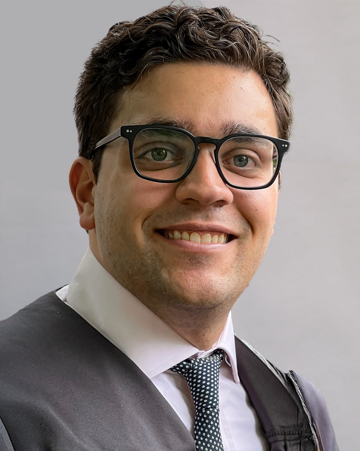 A smiling man with dark, curly hair wearing glasses, a white shirt, a patterned tie, and a dark jacket.