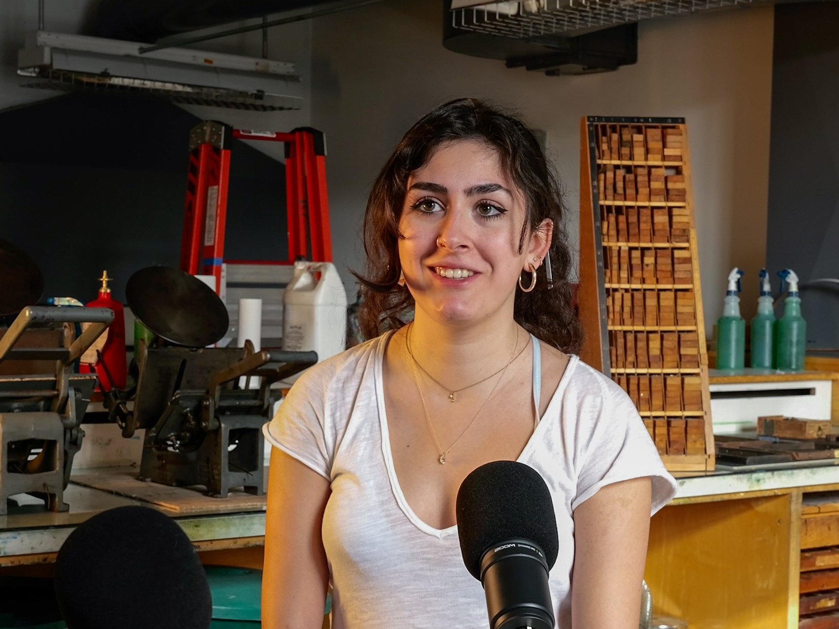 A young woman with dark curly hair and hoop earrings, smiling, sitting in a workshop or studio with woodworking tools and supplies in the background. She is speaking into a microphone.