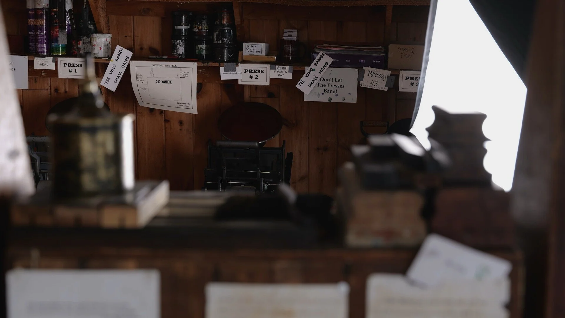 A rustic wooden room with signs and papers attached to the wall, indicating a setup for a shooting or firing range with instructions like 'Press #1' and 'Press #2', and warnings such as 'Yee Who Bangs Shall Hang'. There are some stacked chairs and various supplies visible, suggesting it might be a workshop or training area.
