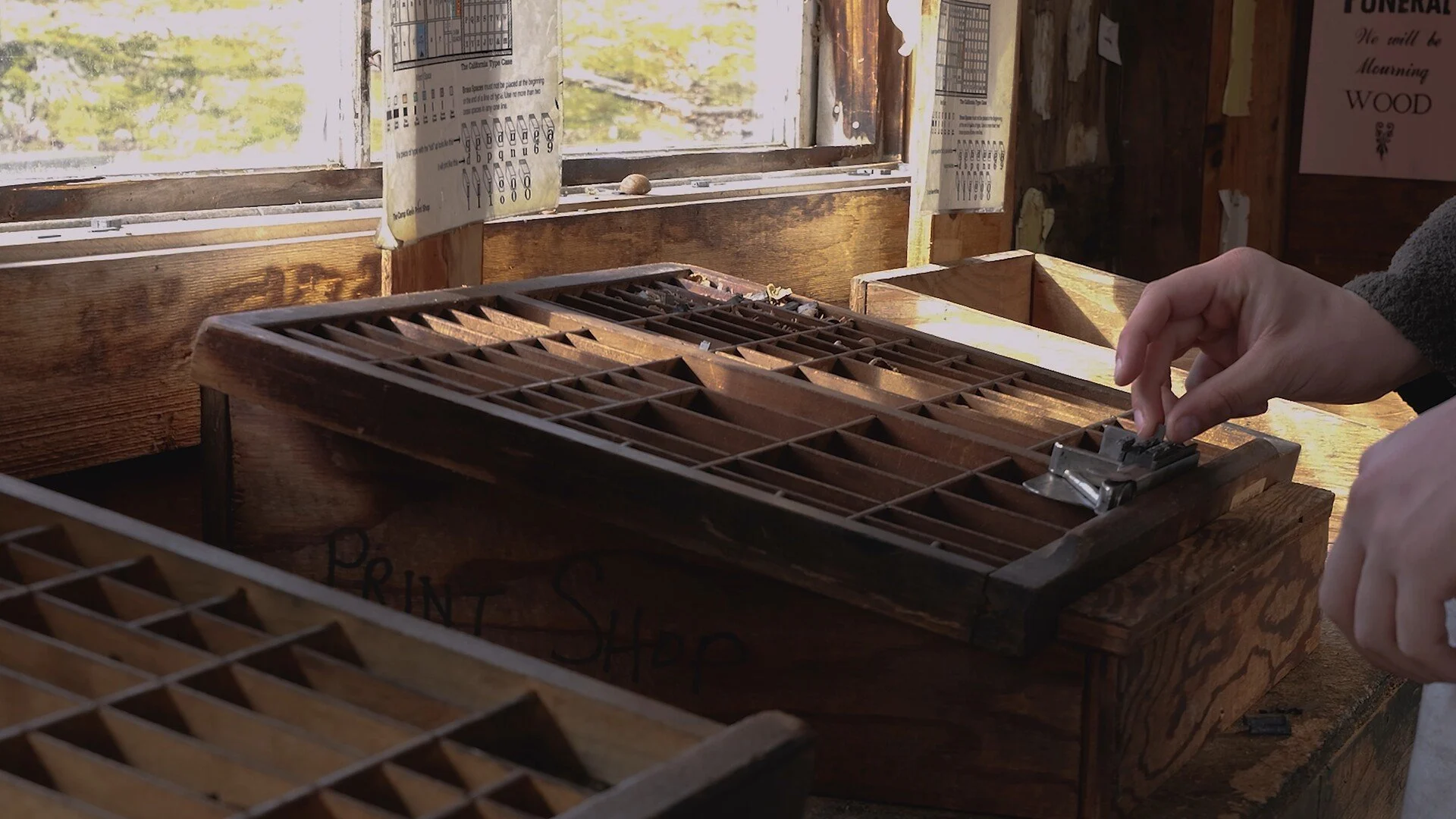 A person using a metal stapler to fasten papers inside a wooden drawer filled with compartments, near a window with sunlight streaming in.