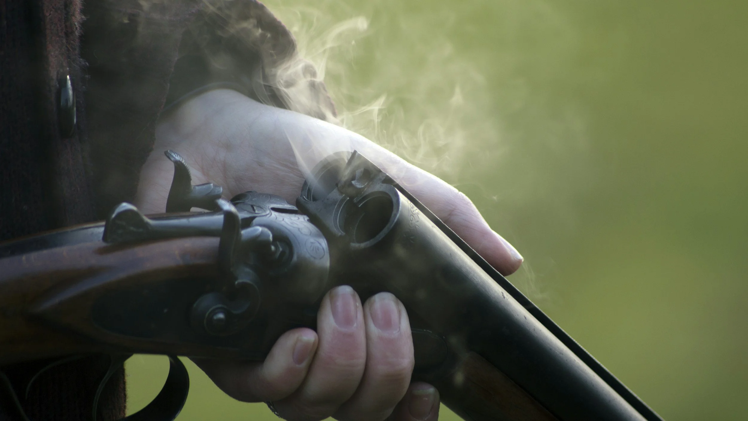 Close-up of a person holding a smoking vintage rifle with a wooden stock, outdoors in a natural setting.