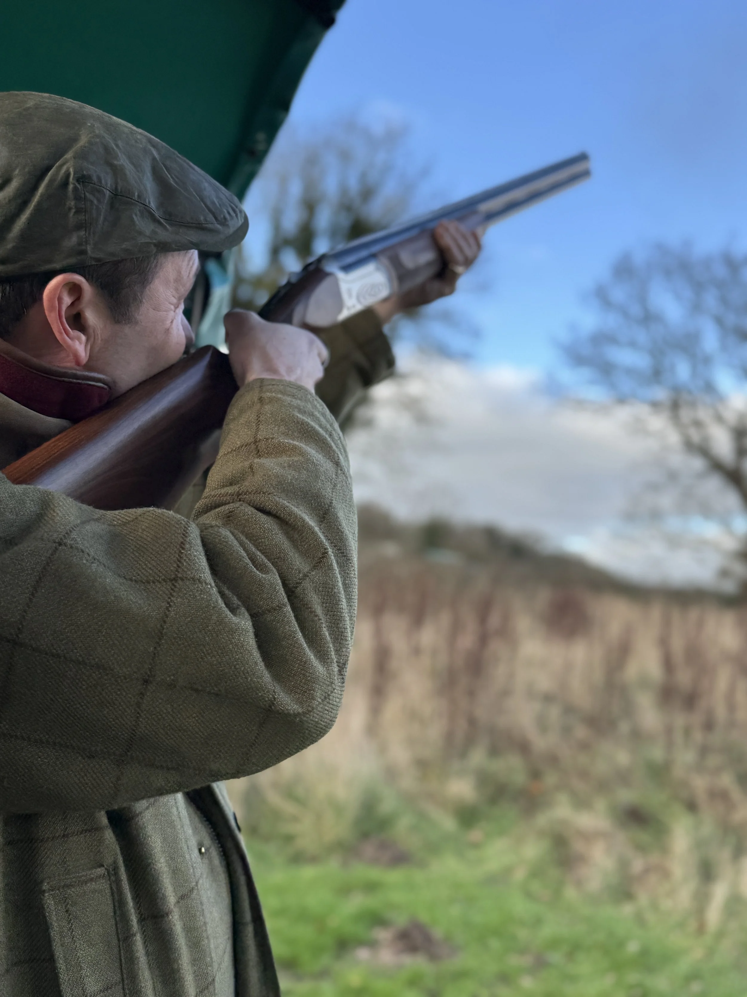 A man aiming a shotgun outdoors, wearing a cap and brown jacket, with trees and blue sky in the background.