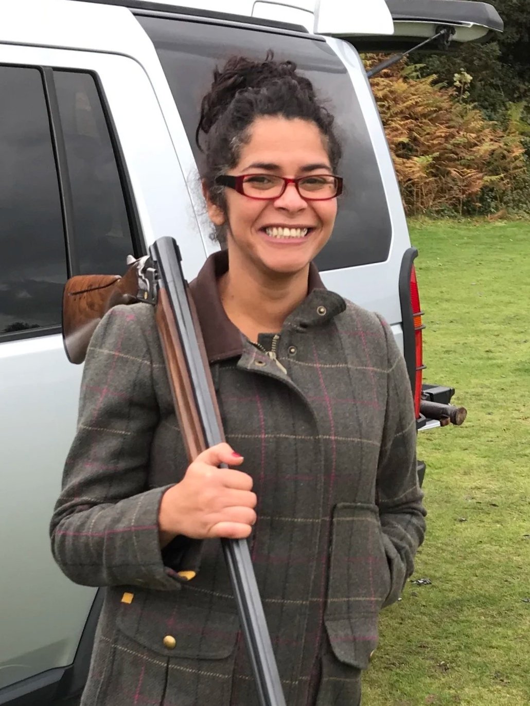 Woman with glasses holding a shotgun, standing outdoors near a vehicle with an open hatchback, and a background of grass and trees.
