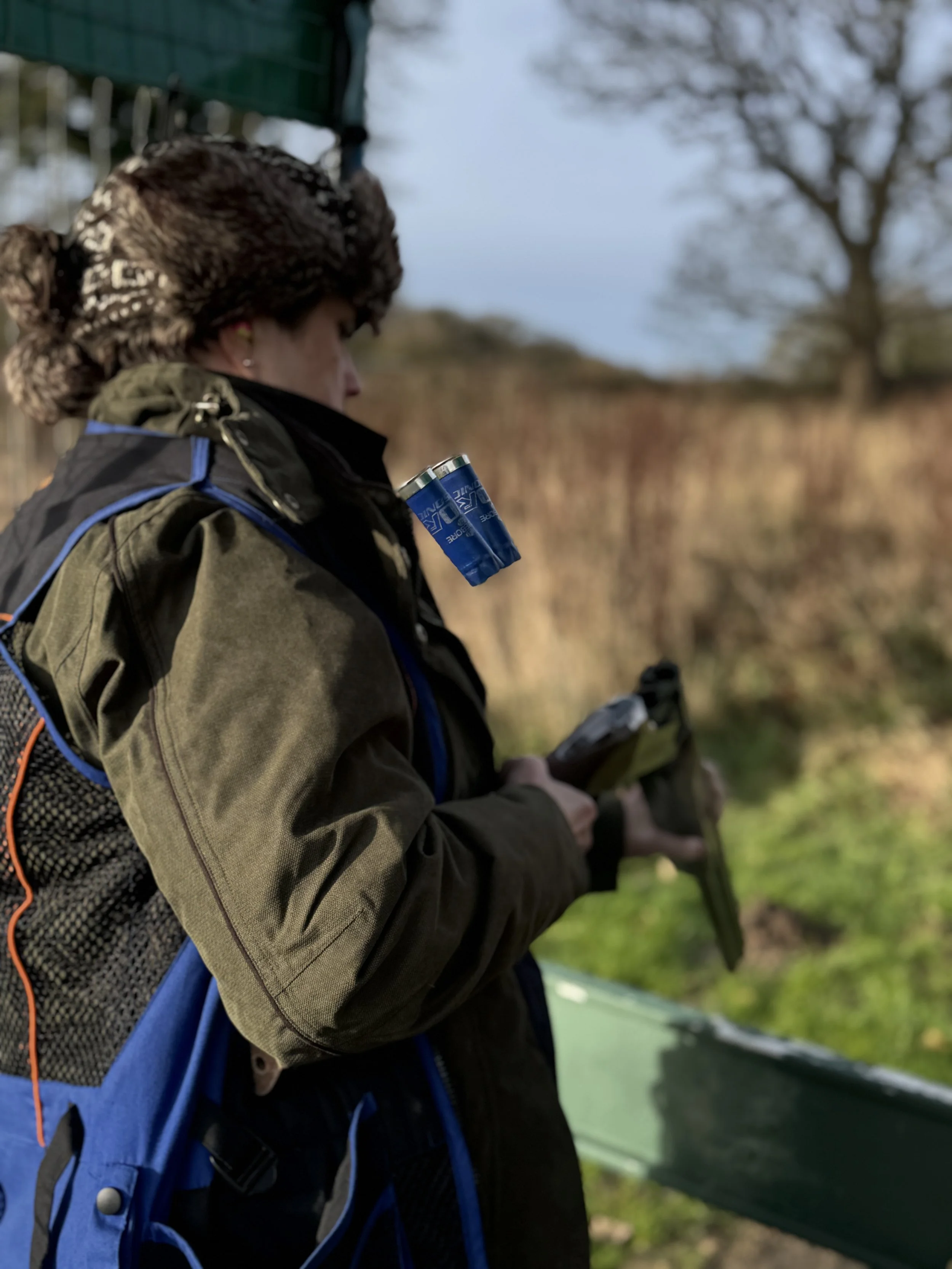 Person standing outdoors, dressed warmly with a backpack and holding a smartphone, with trees and grass in the background.