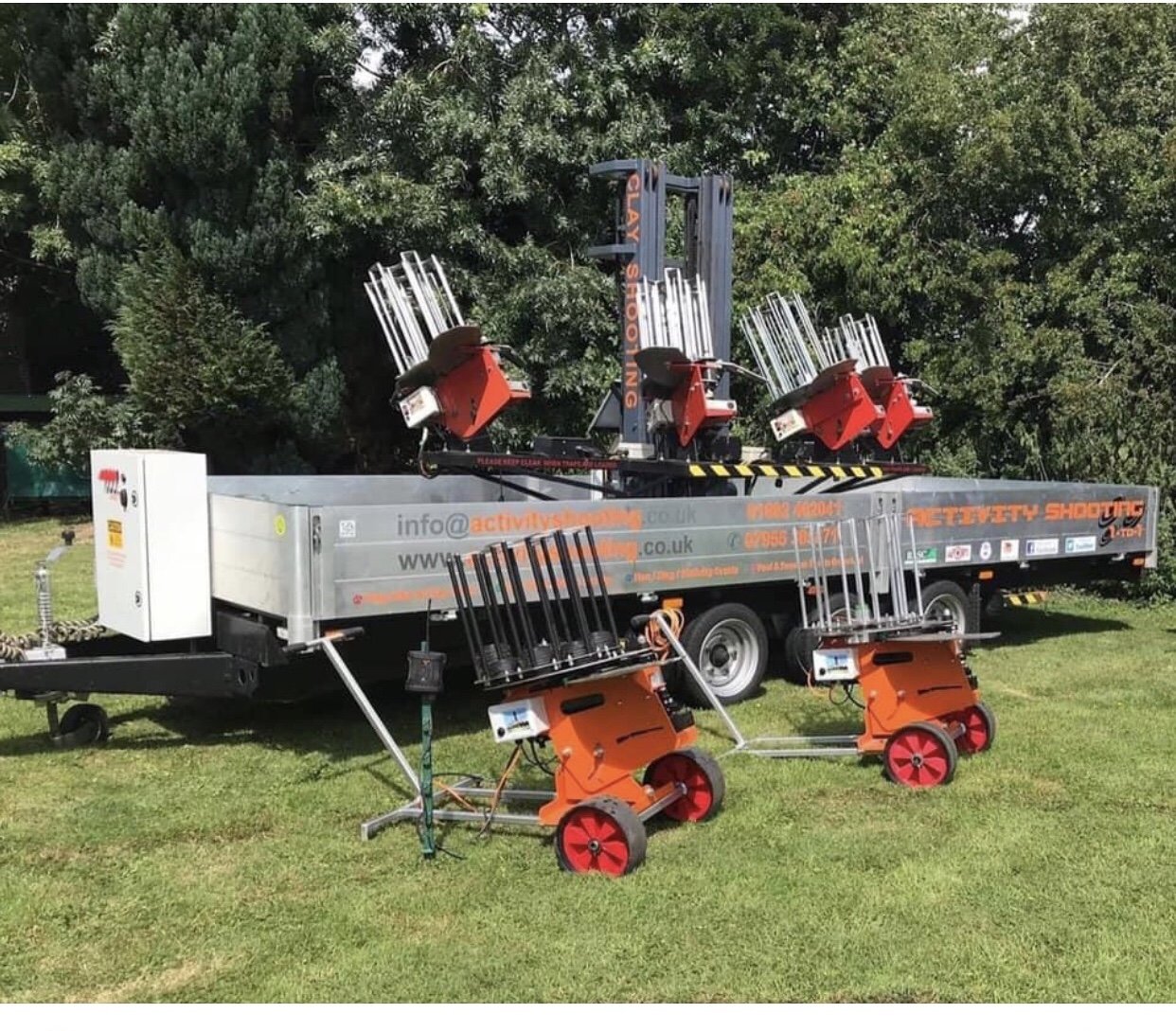 A mobile setup of laser tagging equipment with multiple laser guns on stands, mounted on a trailer, with additional equipment on orange carts, outdoors on grass with trees in the background.