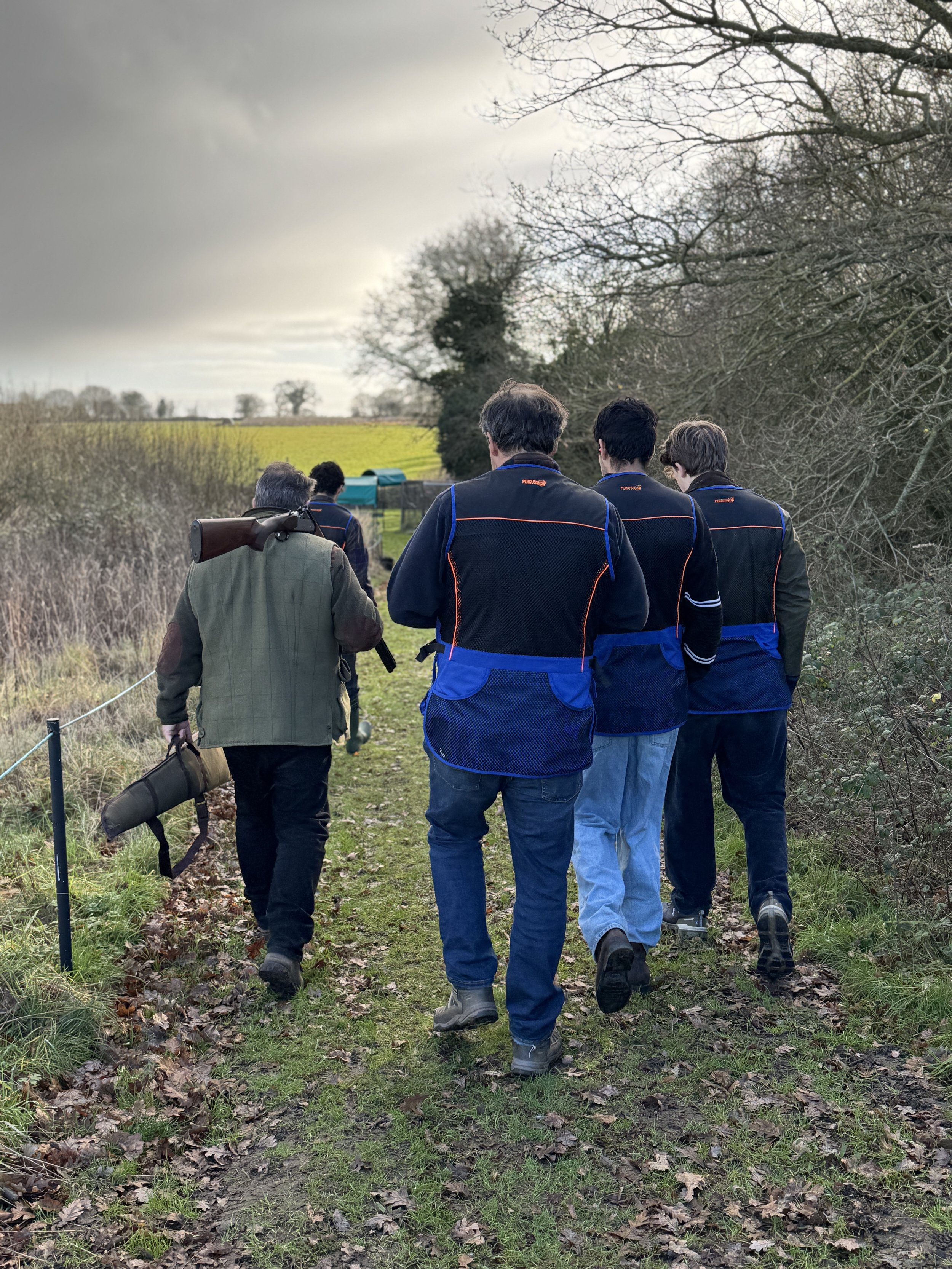 A group of men walking along a dirt trail through a rural area, with grassy fields and leafless trees on either side, under a cloudy sky.