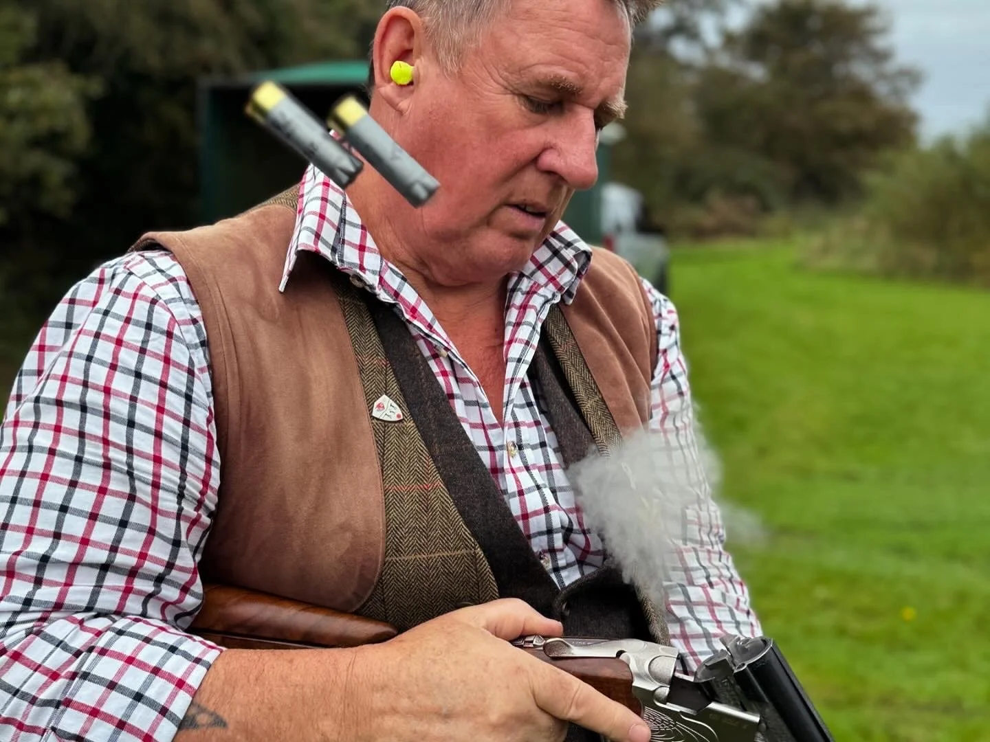 A man wearing a checkered shirt, brown vest, and hearing protection fires a handgun outdoors, with smoke coming from the gun's barrel.