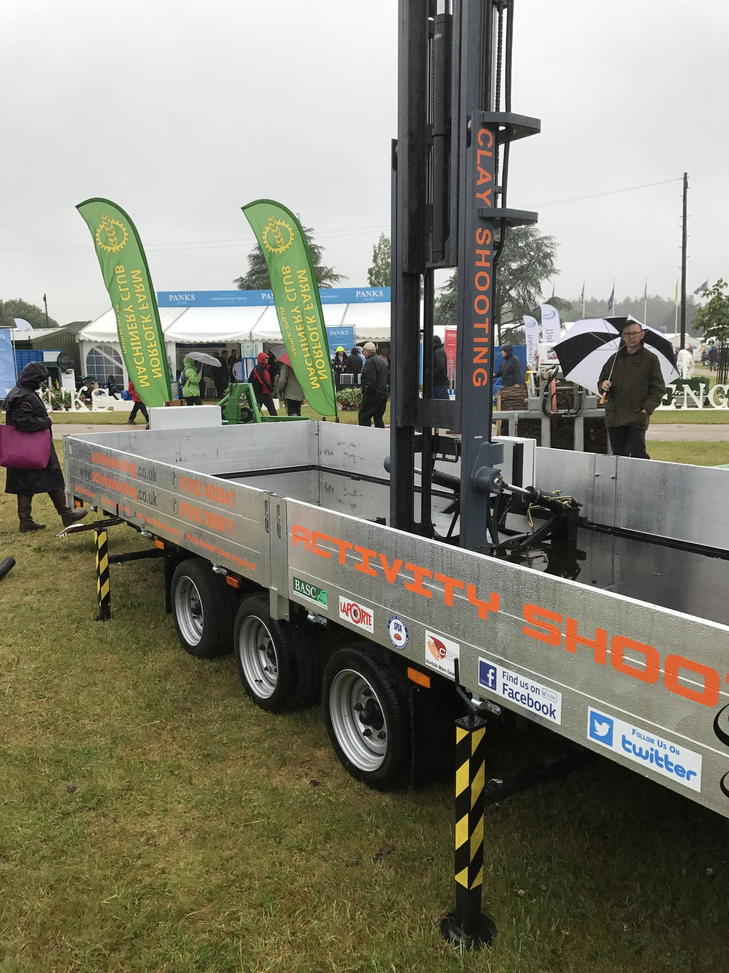 A metal vehicle with a crane-like lifting device labeled 'ACTIVITY SHOOT' at an outdoor event on a cloudy day, with people holding umbrellas in the background.