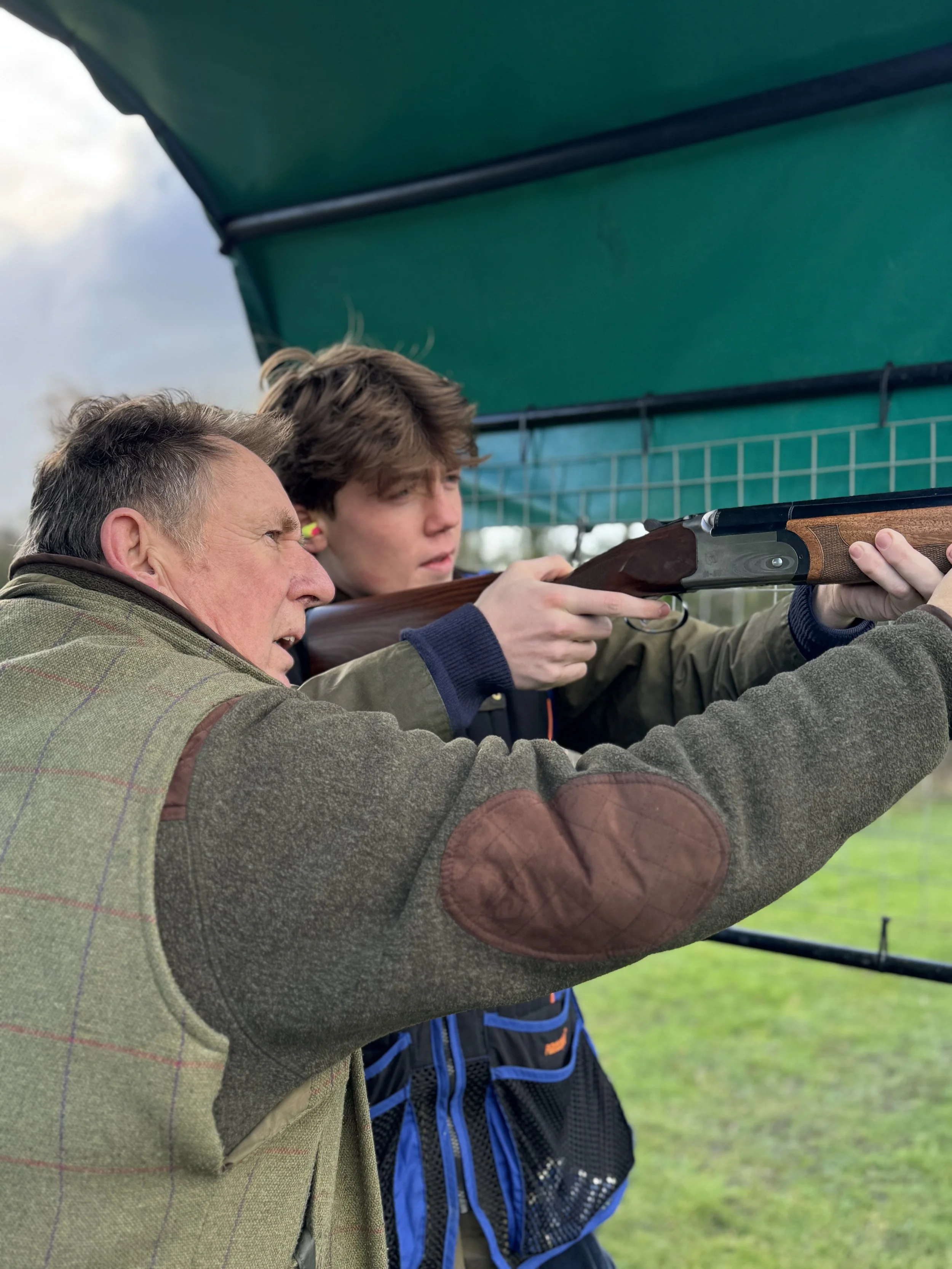 An older man teaching a young boy how to shoot with a rifle outdoors, under a green canopy, on a grassy field.