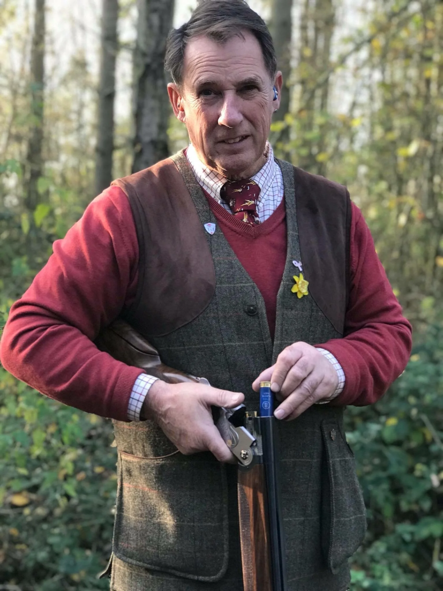 A man dressed in traditional outdoor hunting attire is holding a shotgun, standing in a forest with autumn foliage.