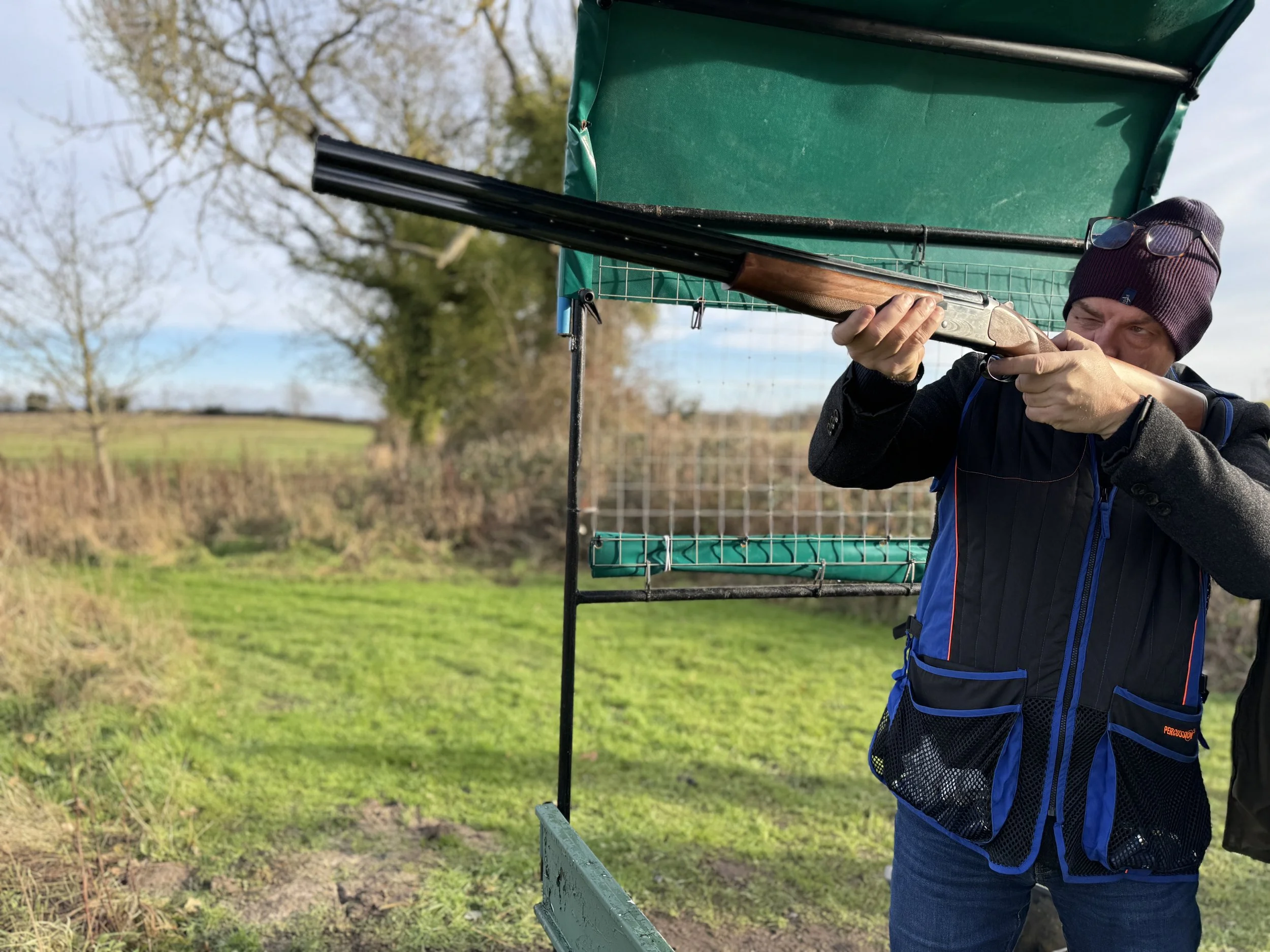 A person aiming a shotgun outdoors near a green hunting blind, with trees and a grassy field in the background.