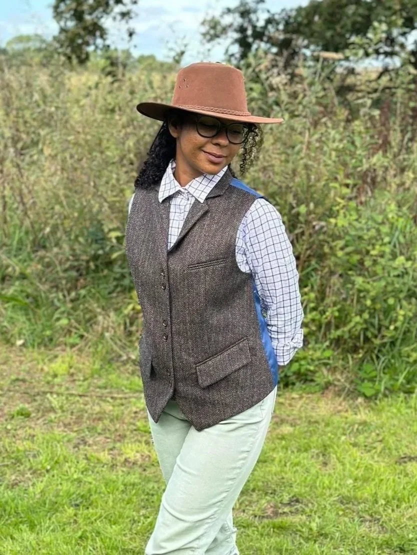 A woman standing outdoors in a grassy area, wearing a brown wide-brimmed hat, black glasses, a checkered shirt, a brown vest, and light-colored pants.