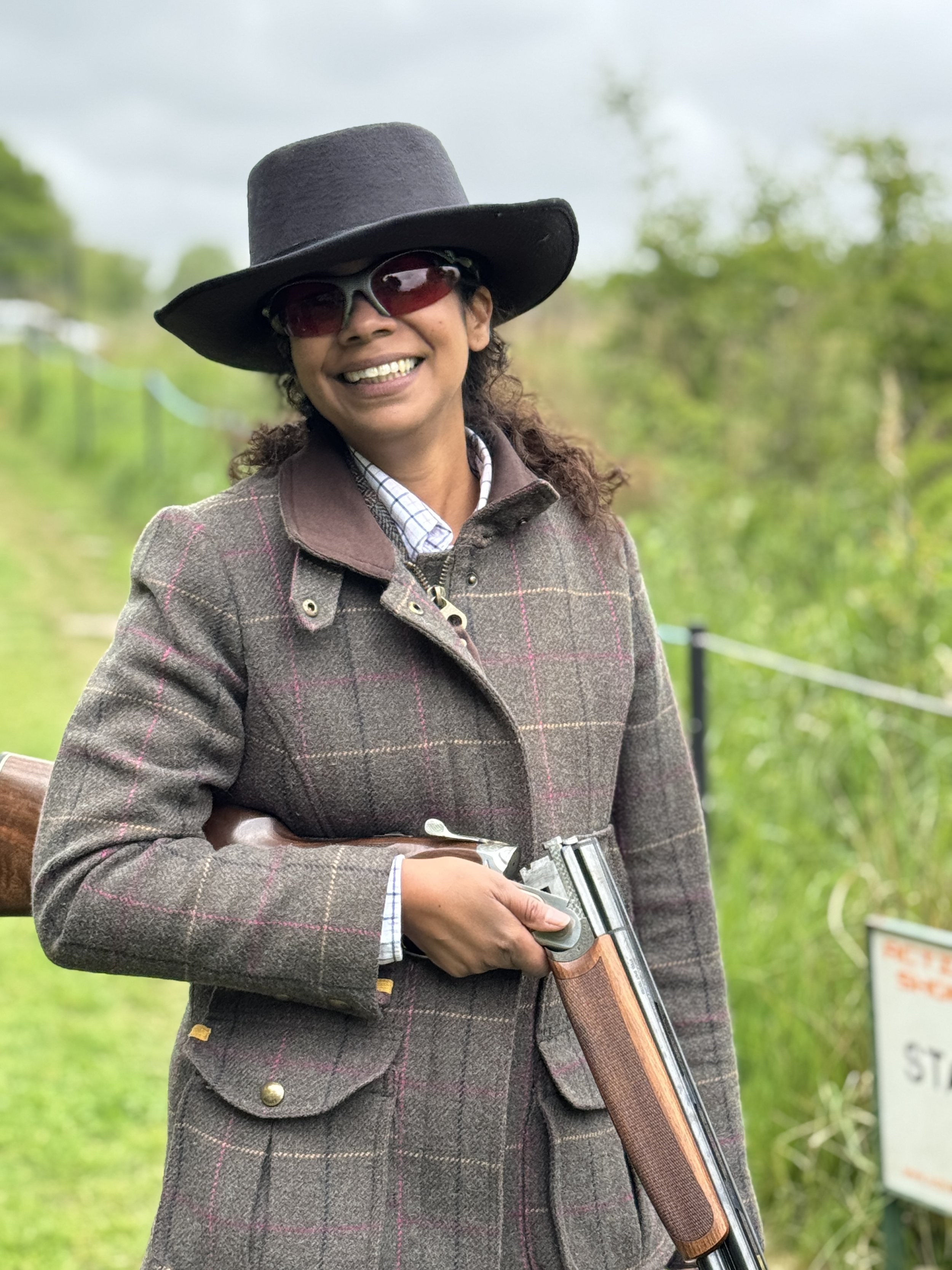 A woman smiling outdoors, wearing a wide-brimmed hat, sunglasses, a plaid jacket, and holding a shotgun.