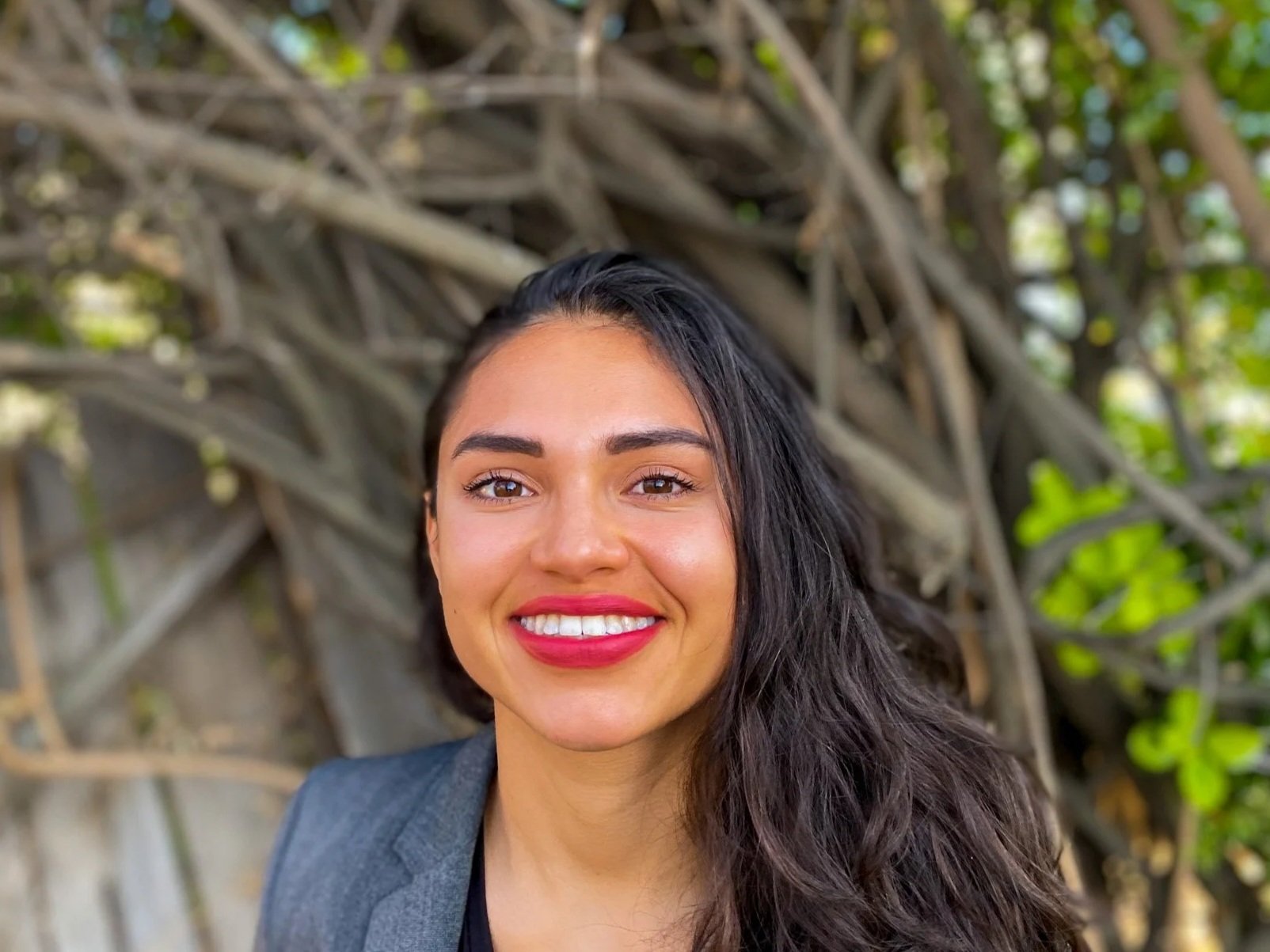 A woman with long dark hair, smiling with red lipstick, wearing a blazer, standing outdoors in front of a background of tree branches and green leaves.
