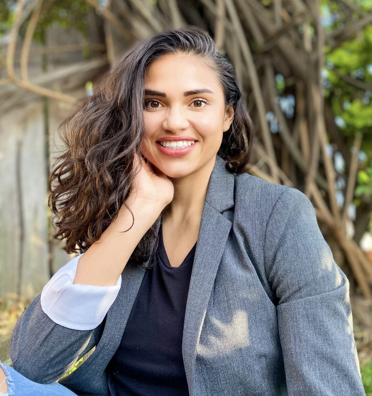 A young woman with long, curly brown hair smiling outdoors, wearing a gray blazer and black shirt, with a background of intertwined tree branches and greenery.