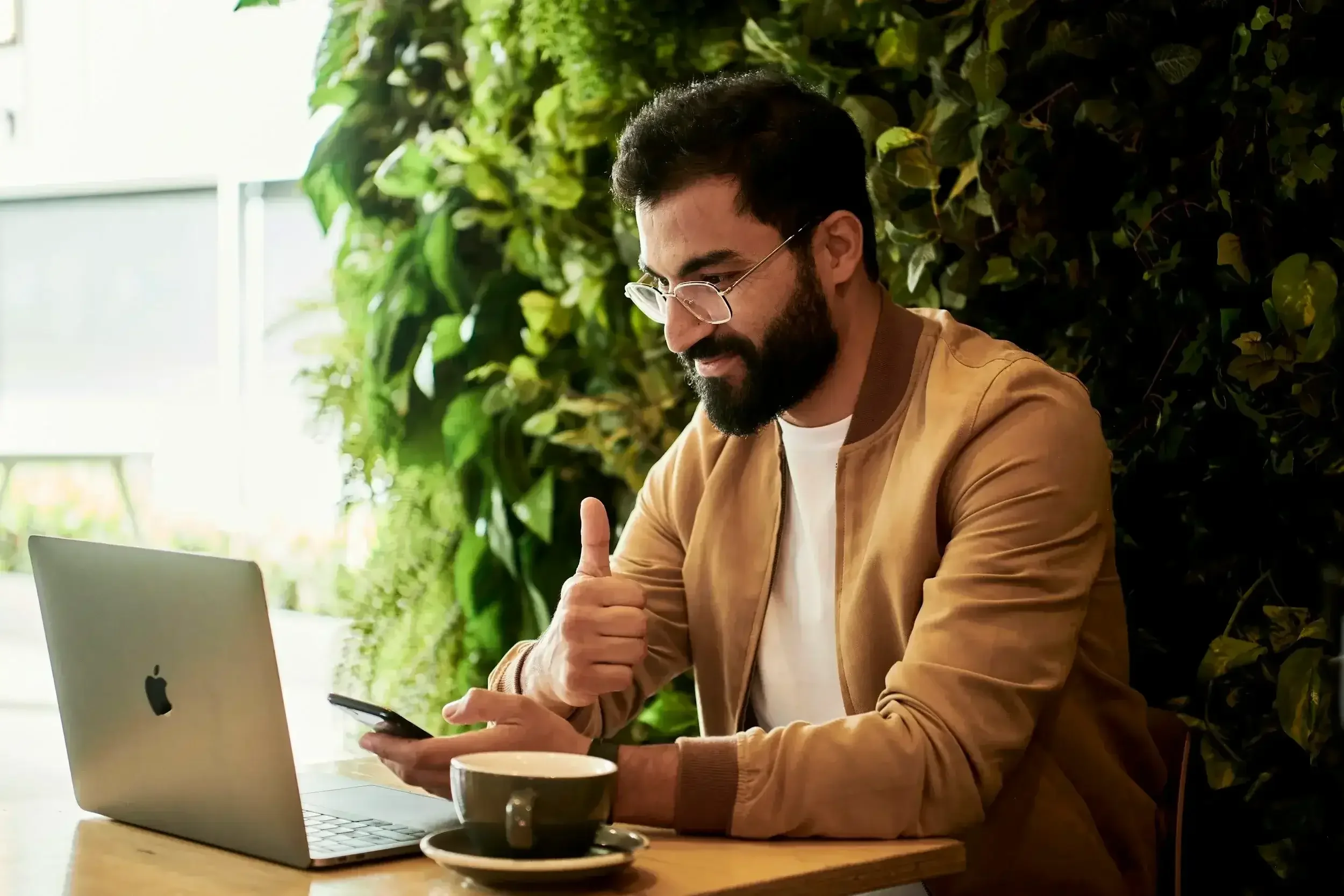 Bearded man with tan coat sitting in front of a laptop with a cup of coffee with a thumbs up