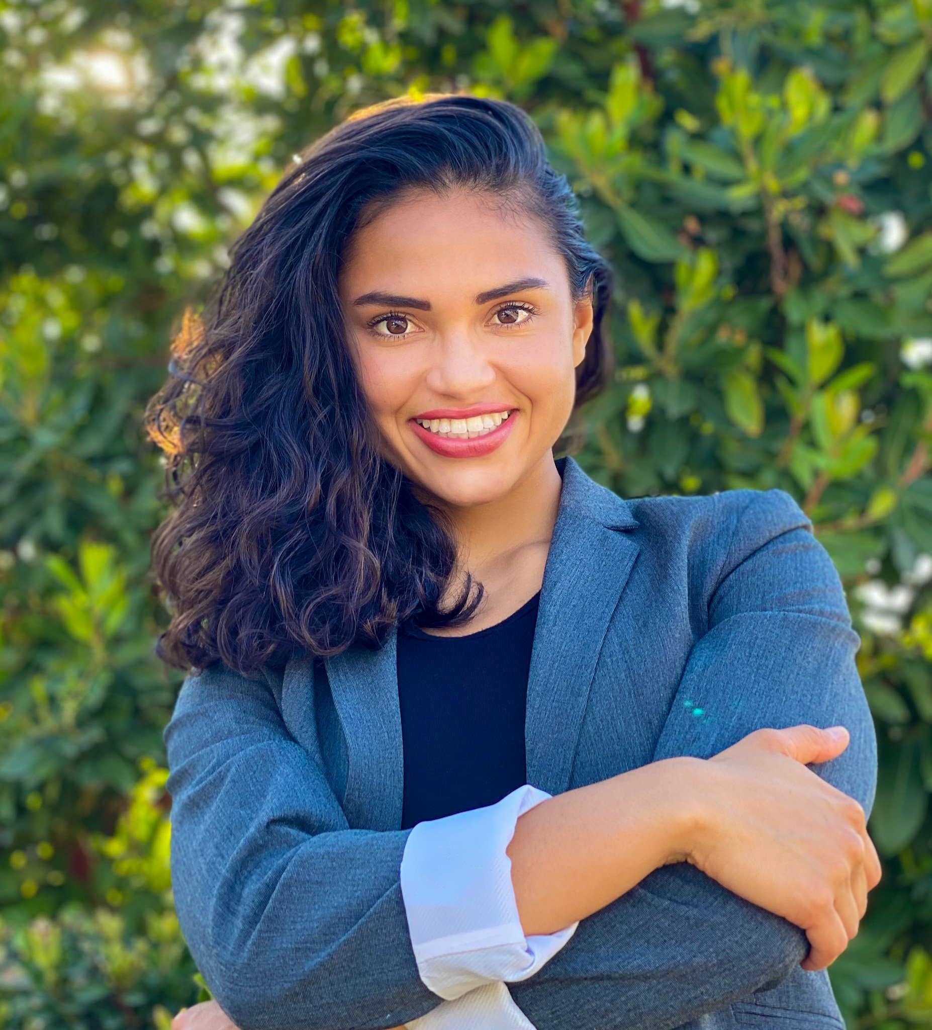 A woman with semi curly dark hair wearing a gray blazer and black shirt stands outdoors in front of greenery, smiling at the camera.