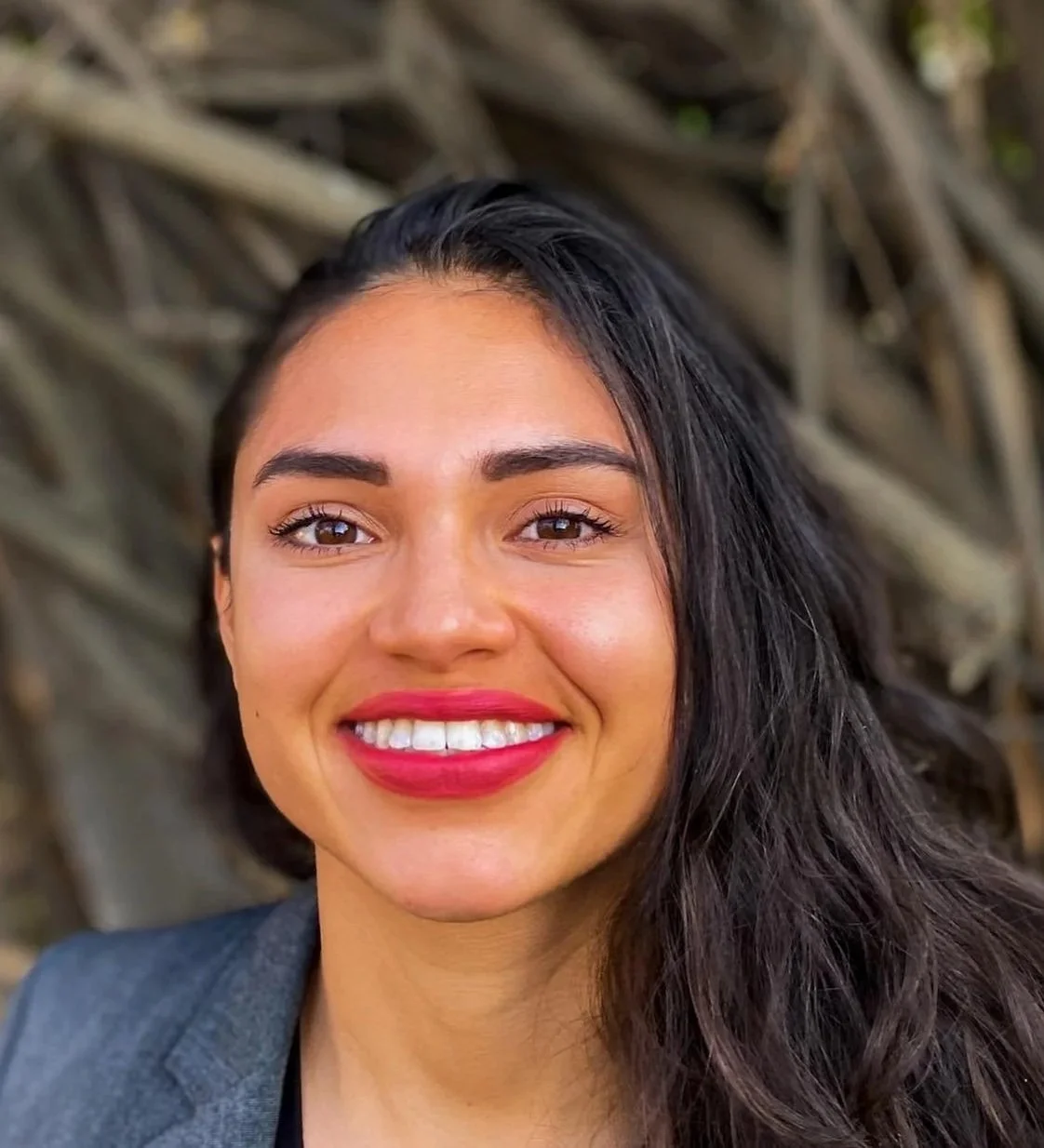 Close-up portrait of a smiling woman with dark hair, wearing a gray blazer, with a blurred natural background.