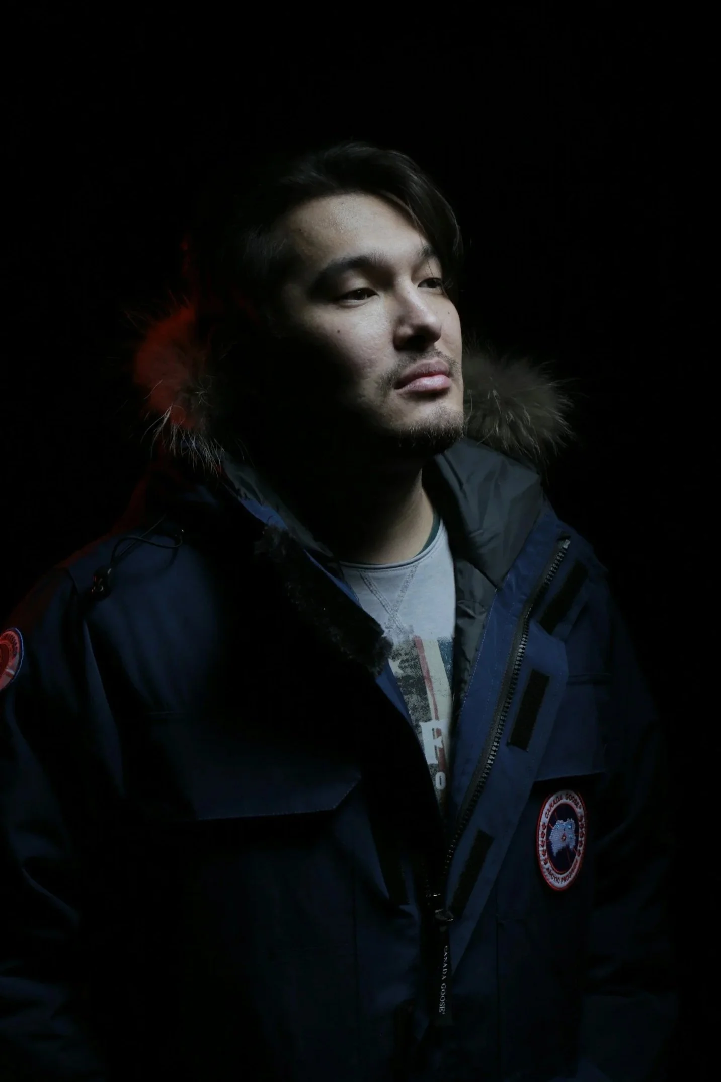A young man with dark hair and facial hair, wearing a dark winter coat with a fur-lined hood, looking to the side against a dark background.