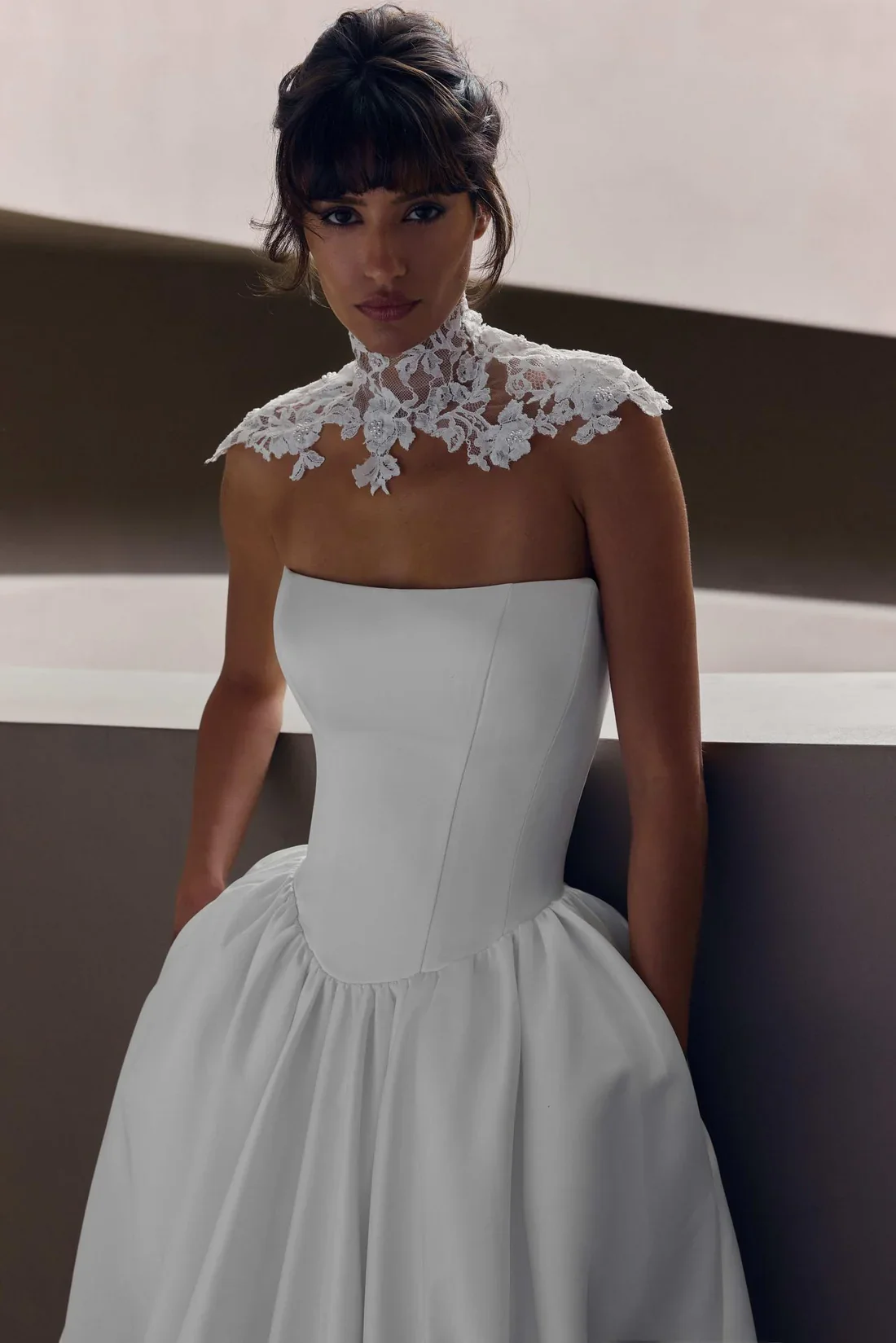 A woman in a white wedding dress with a lace high-neck collar, sitting against a modern, minimalist background.
