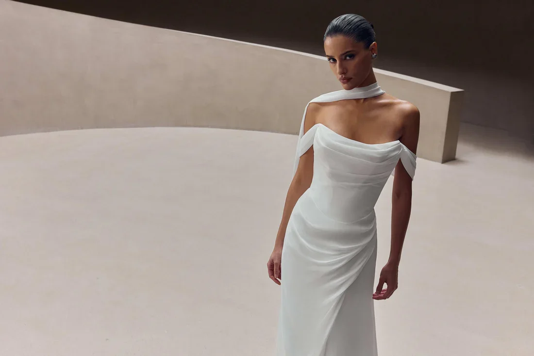 A woman in a sleek, off-the-shoulder white dress with a high slit, standing on a minimalist, curved beige background.