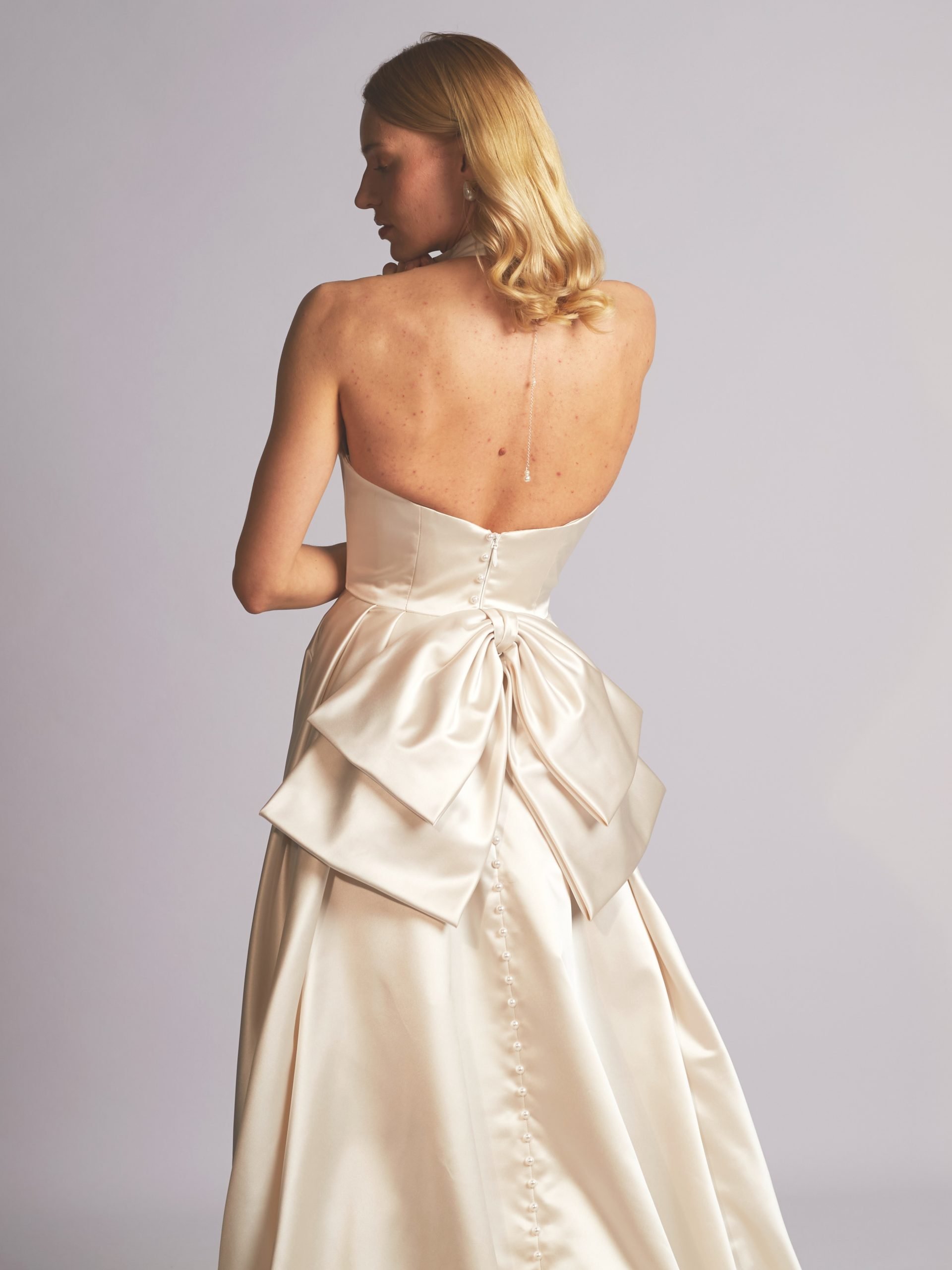A woman in a strapless satin wedding dress with a large bow on the back, standing against a plain light gray background.