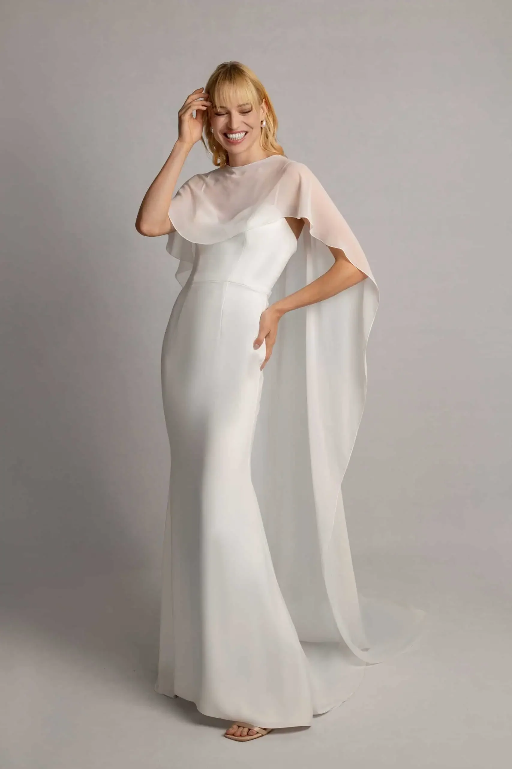A woman in a white wedding dress with a sheer cape, smiling and touching her hair, standing against a plain background.