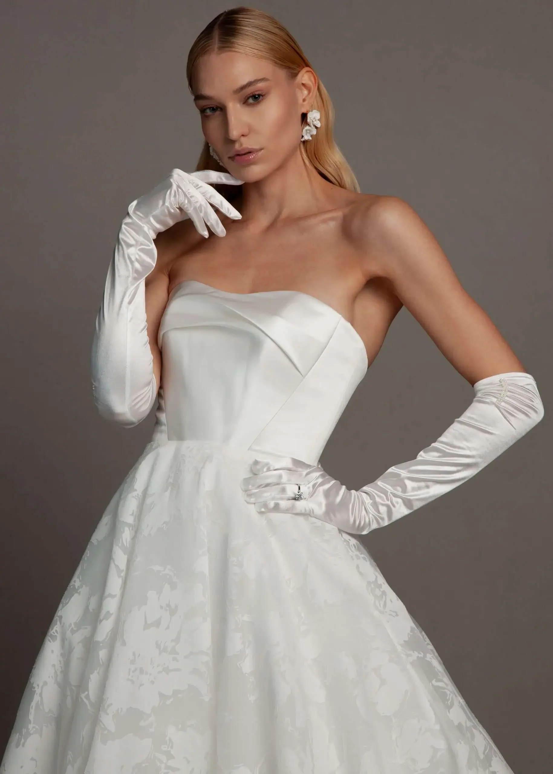 A woman with blonde hair in a white strapless evening gown, wearing long white satin gloves and floral earrings, posing against a plain grey background.